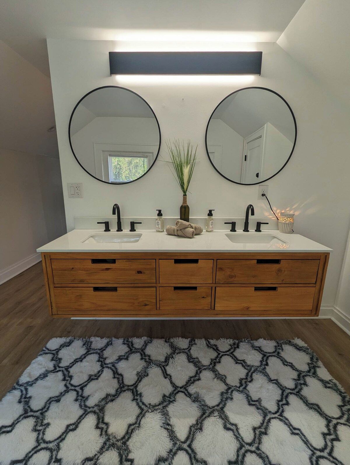 A bathroom with two round mirrors, a white vanity with a wooden base, and a patterned rug on the floor.
