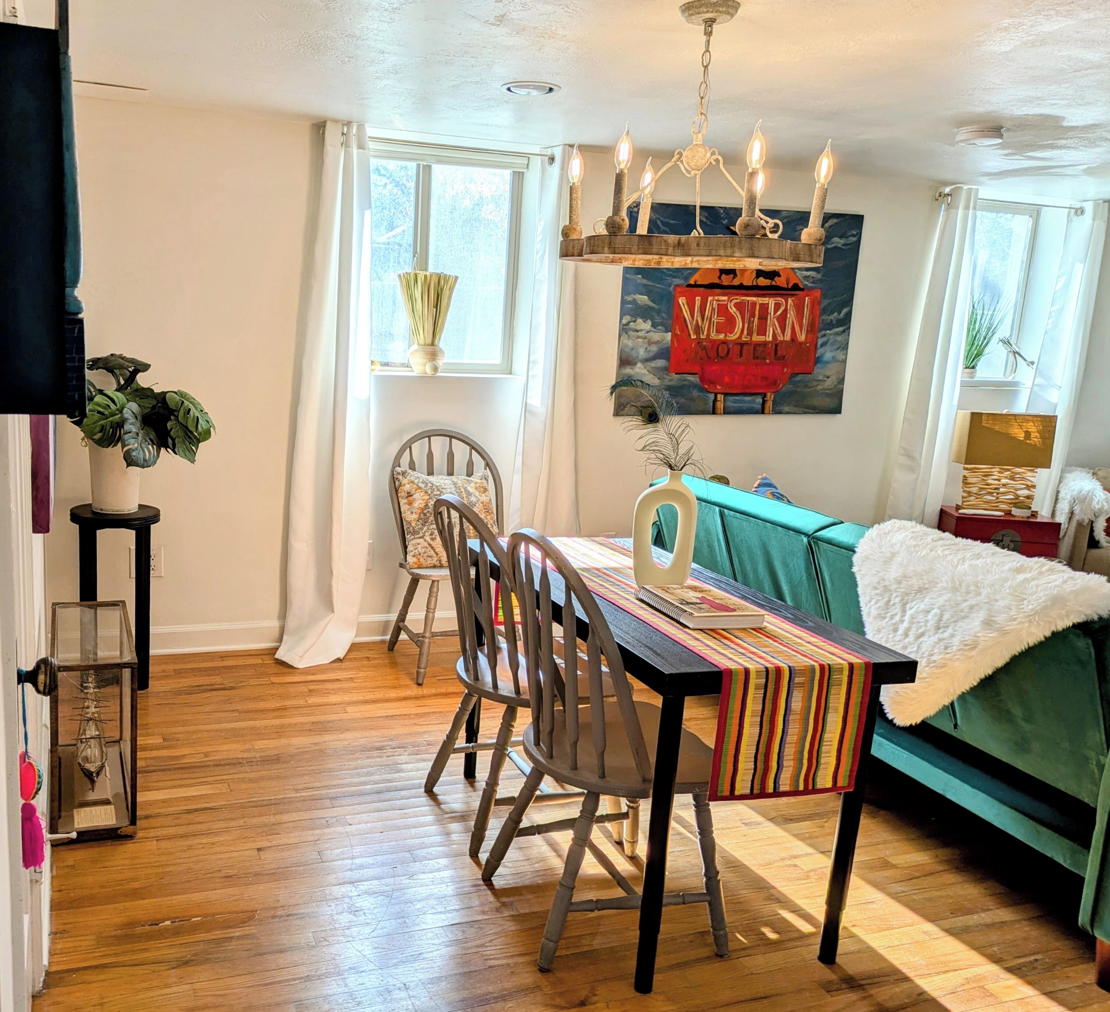 A dining area with a wooden table, chairs, and a chandelier. There is a sofa and a painting of a Western-themed sign in the background.