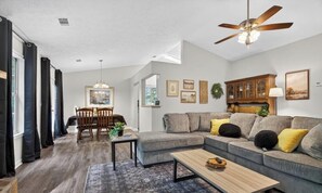 A living room with a sectional sofa, a coffee table, a dining area with chairs, and a ceiling fan.