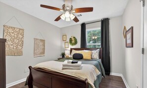 Une chambre à coucher comprenant un lit, un ventilateur de plafond, une fenêtre avec des rideaux et des éléments de décoration murale.