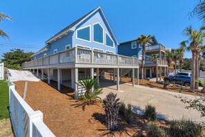 Ein zweigeschossiges Strandhaus mit Balkon, überdachter Auffahrt und einer sandigen Fläche mit Pflanzen.