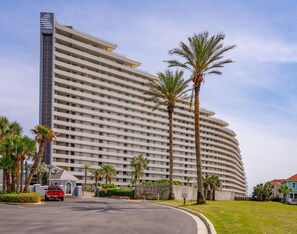 Un hotel de diverses plantes amb façana corba, palmeres i un vehicle vermell estacionat al davant.