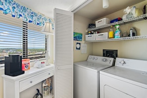 A laundry room with a washing machine, dryer, and shelves with cleaning supplies. - 🌴Edgewater #1400 | BeachFront 2BR condo | Pool 🏖️ (Miramar Beach)