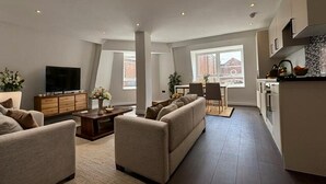 Ein modernes Wohnzimmer mit einer Sofagarnitur, einem Couchtisch und einem Fernseher. Es gibt einen Essbereich mit Tisch und Stühlen sowie eine Küche mit Schränken und einer Arbeitsplatte.