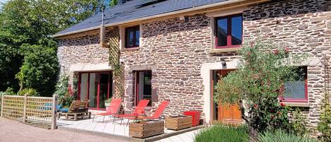 A stone building with a sloped roof, a covered patio with red chairs, and a wooden fence.