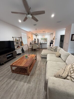 A modern living room with a flat-screen TV, wooden coffee table, beige sofa, and a ceiling fan. - Beautiful pet friendly three bedroom home with Pool near beach and attraction's (St. Augustine)