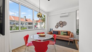 Ein modernes Wohnzimmer mit einem runden Esstisch, roten Stühlen, einer Couch und Blick auf Gebäude durch große Fenster.