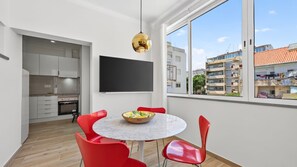 A modern kitchen with white cabinets and a built-in oven, a dining area with a round table and red chairs, and a large window with a view of a residential building. - T2 Moderno c/Varanda em Portimão by Lovelystay (Portimão)