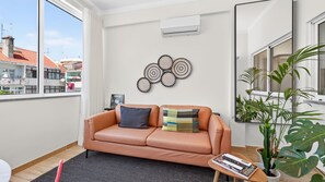 A modern living room with a leather sofa, a coffee table, a wall-mounted air conditioner, and a large window with a view of buildings. - T2 Moderno c/Varanda em Portimão by Lovelystay (Portimão)