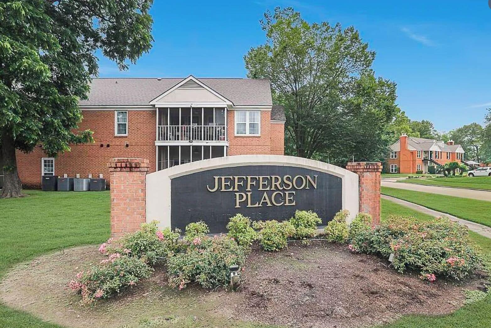A brick building with a sign reading 'JEFFERSON PLACE' surrounded by a well-maintained lawn and flowering shrubs.