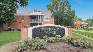 A brick building with a sign reading 'JEFFERSON PLACE' surrounded by a well-maintained lawn and flowering shrubs.