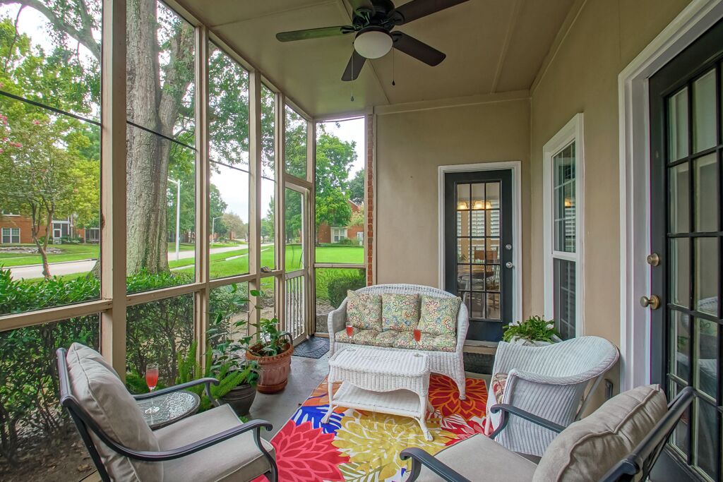 A covered porch with wicker furniture, large windows, and a view of a green landscape.