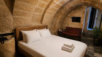 Un dormitorio con techo abovedado de piedra, una cama de madera, una mesita con una silla y una planta en maceta.