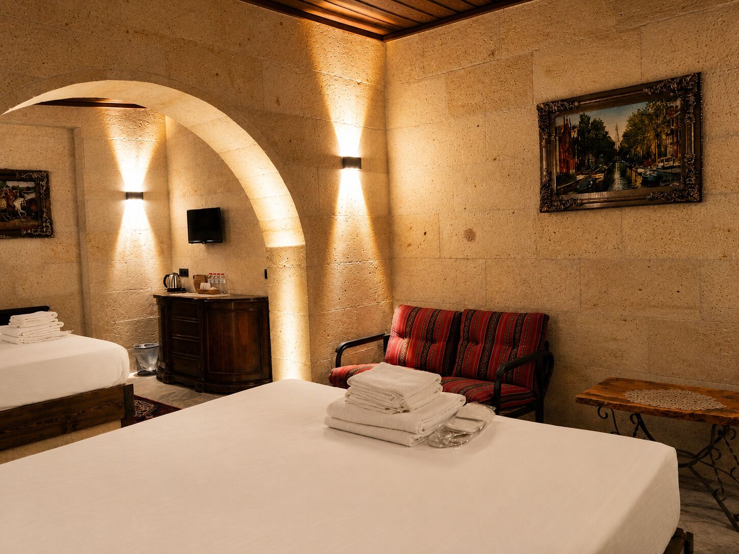 A hotel room with two beds, a red chair, a small table, and a painting on the wall.