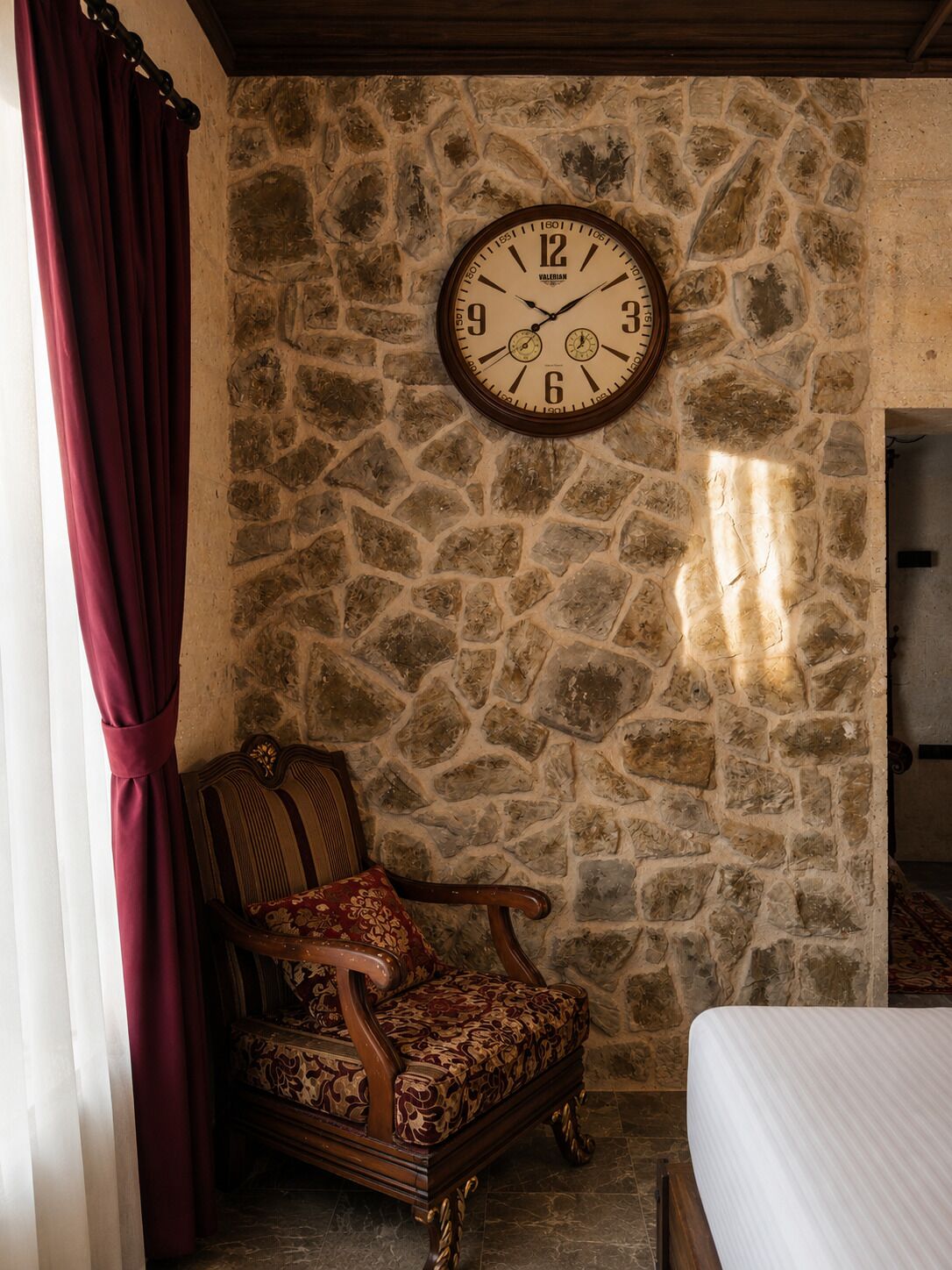 Una habitación con pared de piedra, una silla de madera, una cama y un reloj en la pared.