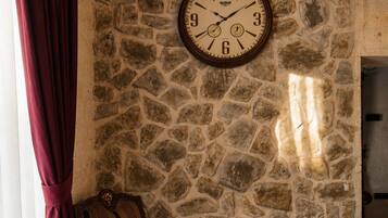 Una habitación con pared de piedra, una silla de madera, una cama y un reloj en la pared.