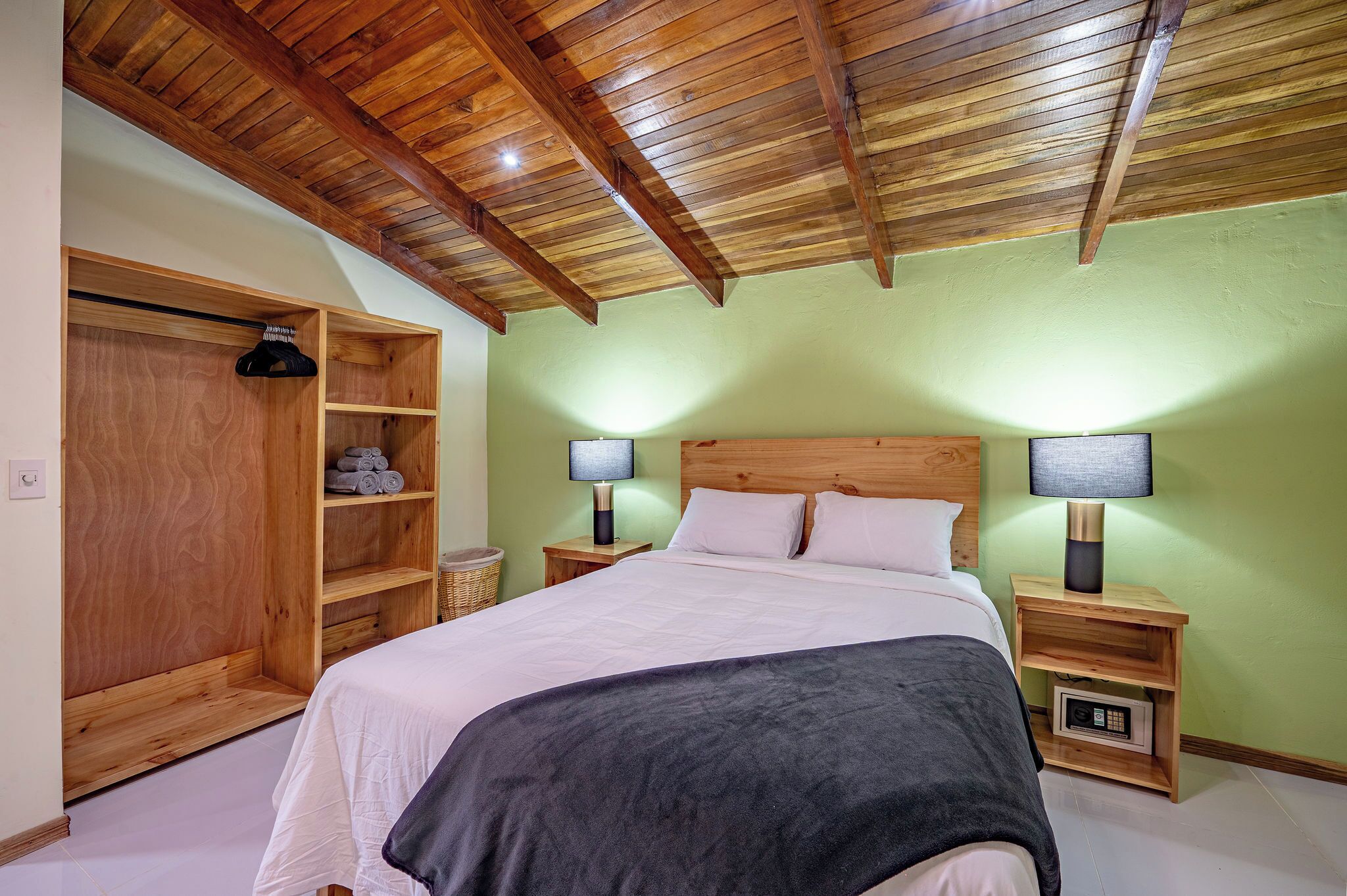 Ein Schlafzimmer mit Holzbalkendecke, einem Bett mit weißer und grauer Bettwäsche, zwei Nachttischlampen, einem Holzschrank und einer grünen Wand.