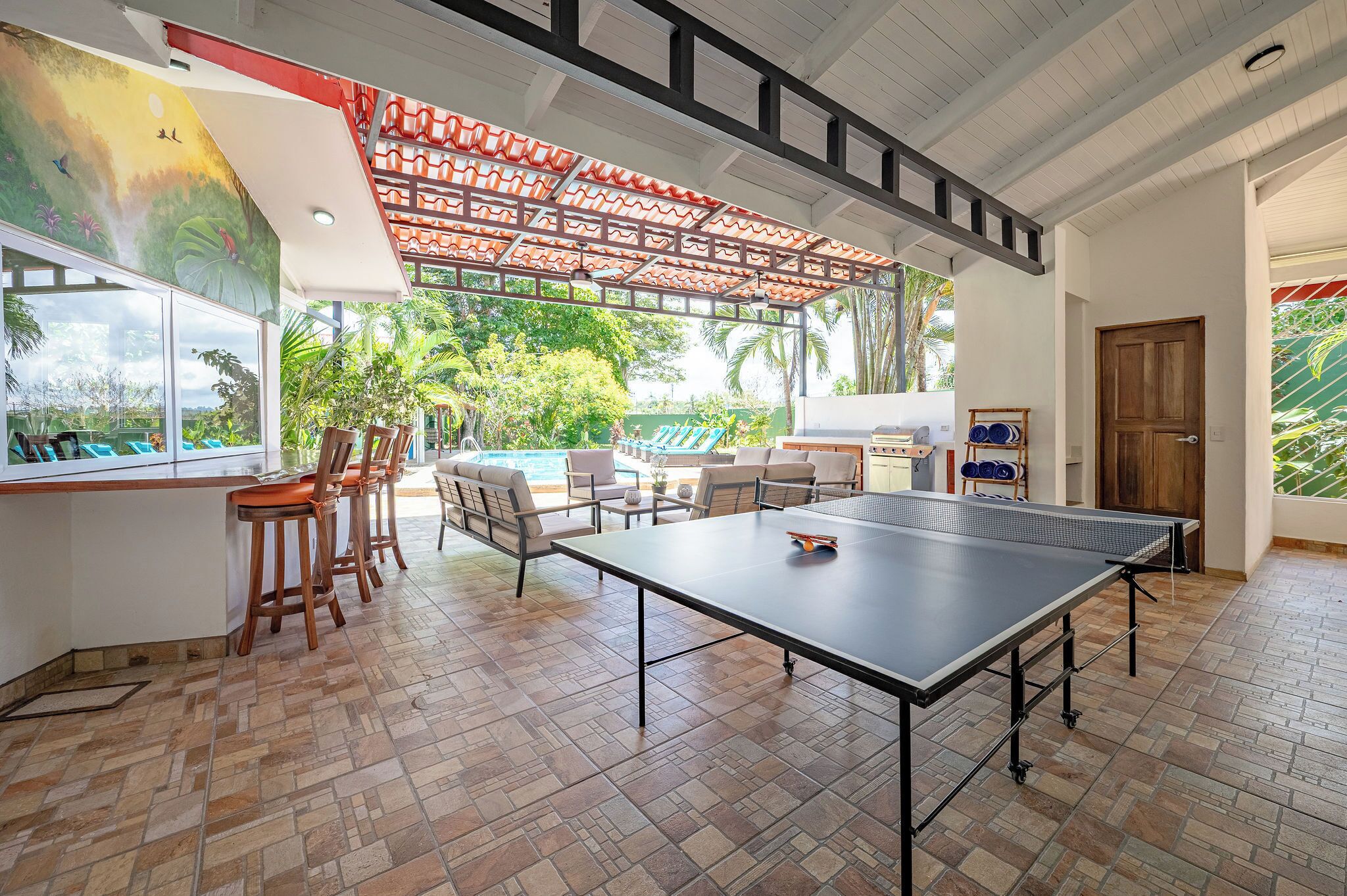 Ein Zimmer mit einem Tischtennis-Tisch, einem Essbereich mit Stühlen, einem Pool und Blick ins Freie.
