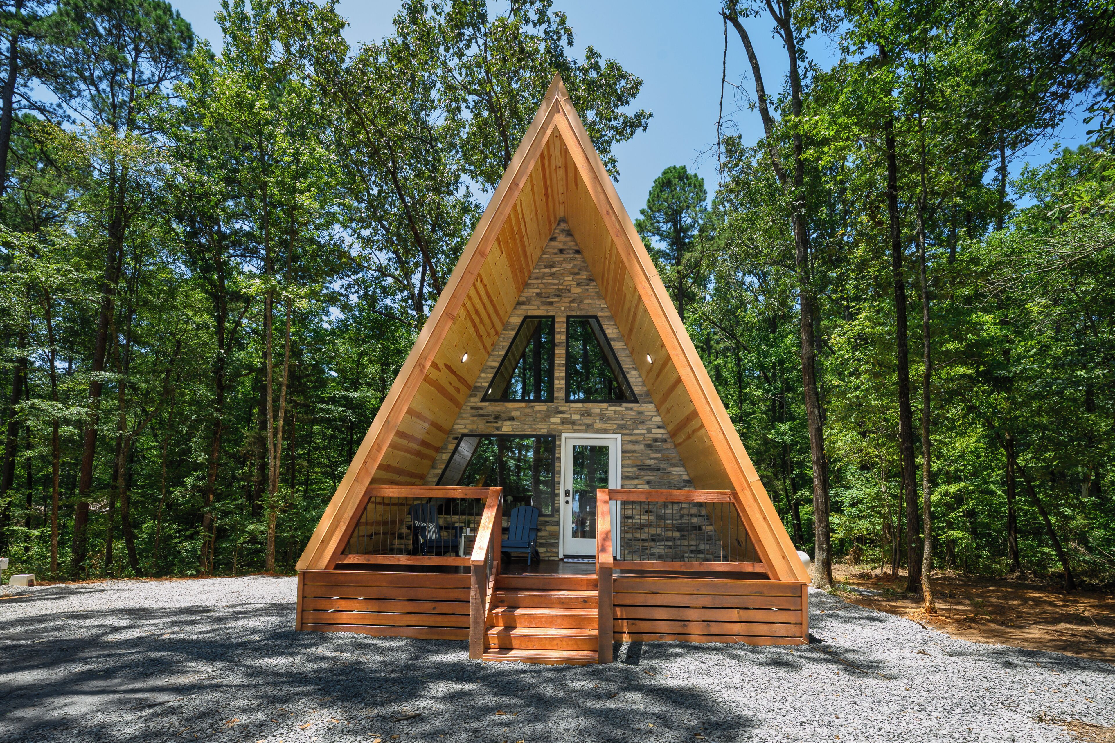 A triangular wooden cabin with a stone facade, a front door, and a set of stairs leading to a porch with two chairs.