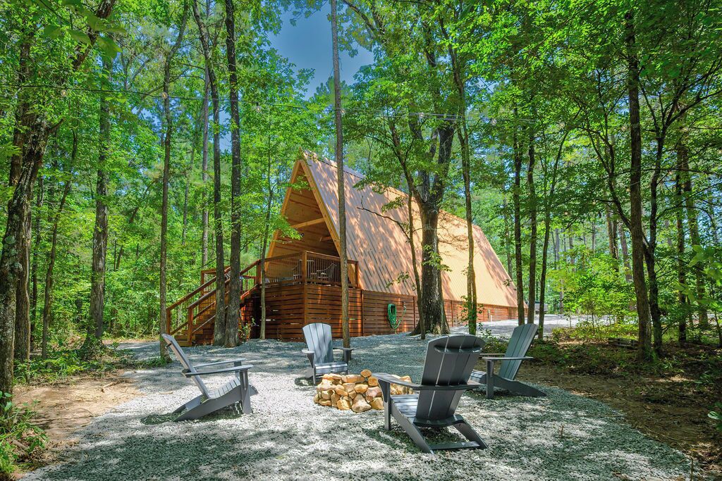 A rustic cabin with a sloped roof, surrounded by a forest. There are outdoor seating areas with chairs and a fire pit.