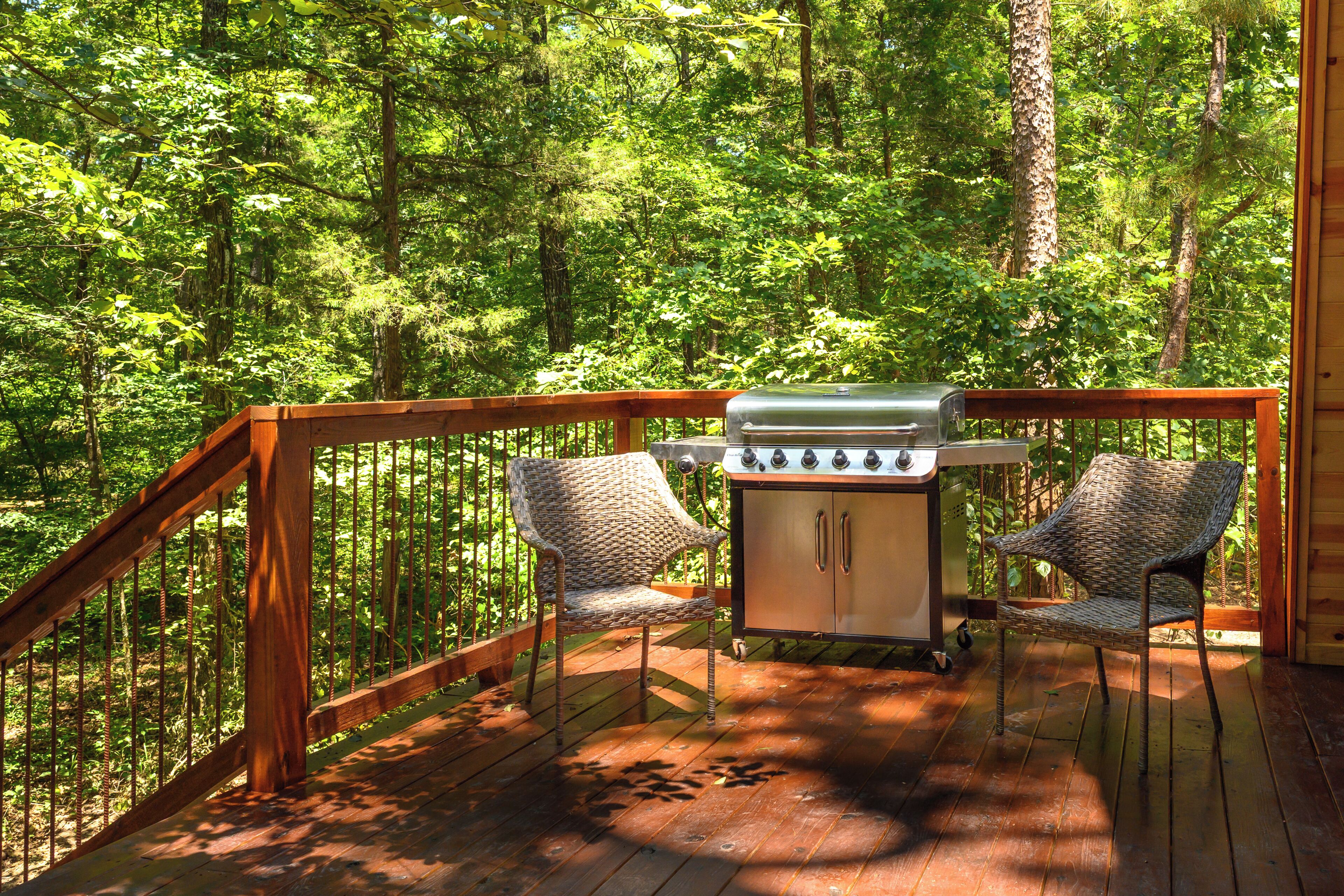 A deck with a grill and two wicker chairs, surrounded by a forest.