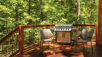 A deck with a grill and two wicker chairs, surrounded by a forest.
