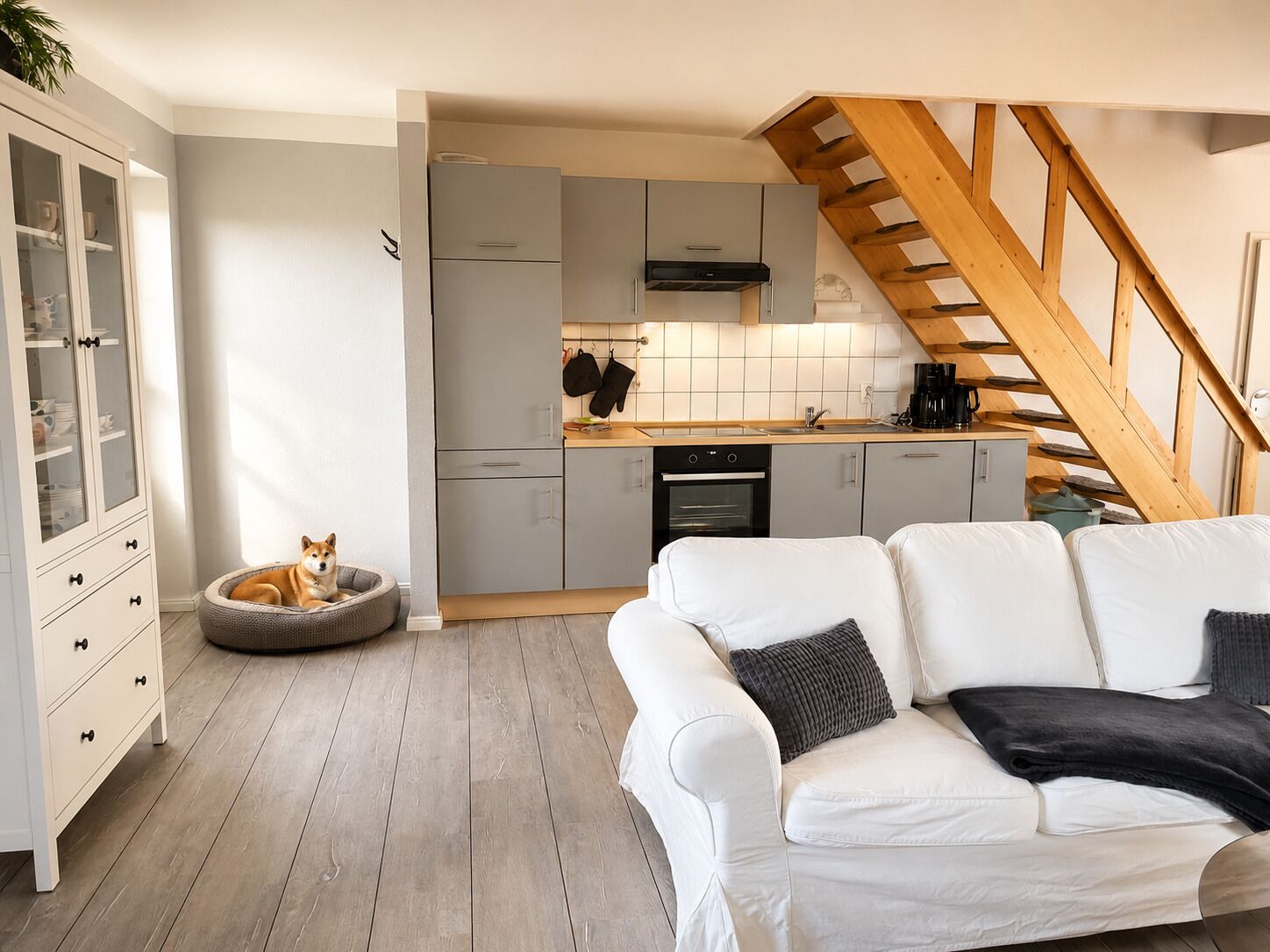 A modern kitchen with grey cabinets, a wooden staircase, and a white sofa with cushions.