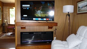A living room with a flat-screen TV mounted on a wooden stand, a fireplace below, and a white armchair.