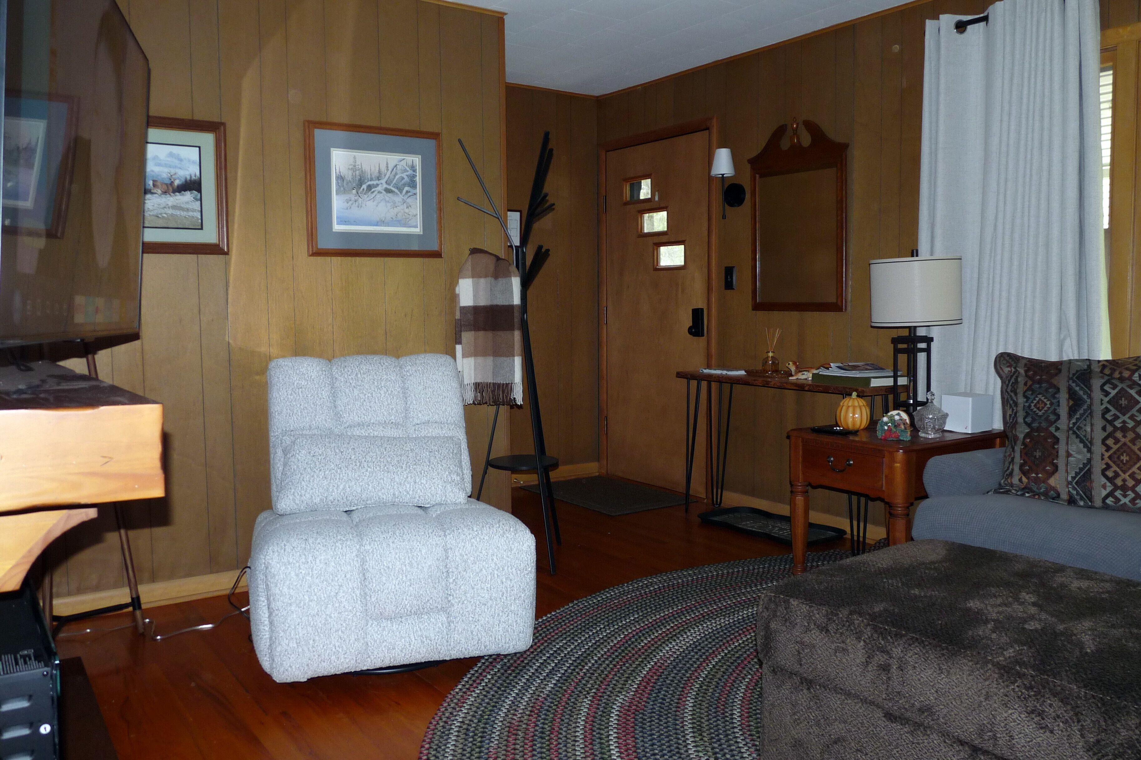 A living room with a grey armchair, a wooden sideboard, a small table with a lamp, and a patterned sofa.