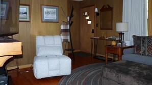 Ett vardagsrum med en grå fåtölj, en träskommod, ett litet bord med en lampa och en mönstrad soffa.