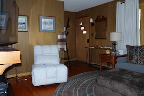 Una sala de estar con un sillón gris, un aparador de madera, una mesita con una lámpara y un sofá estampado.