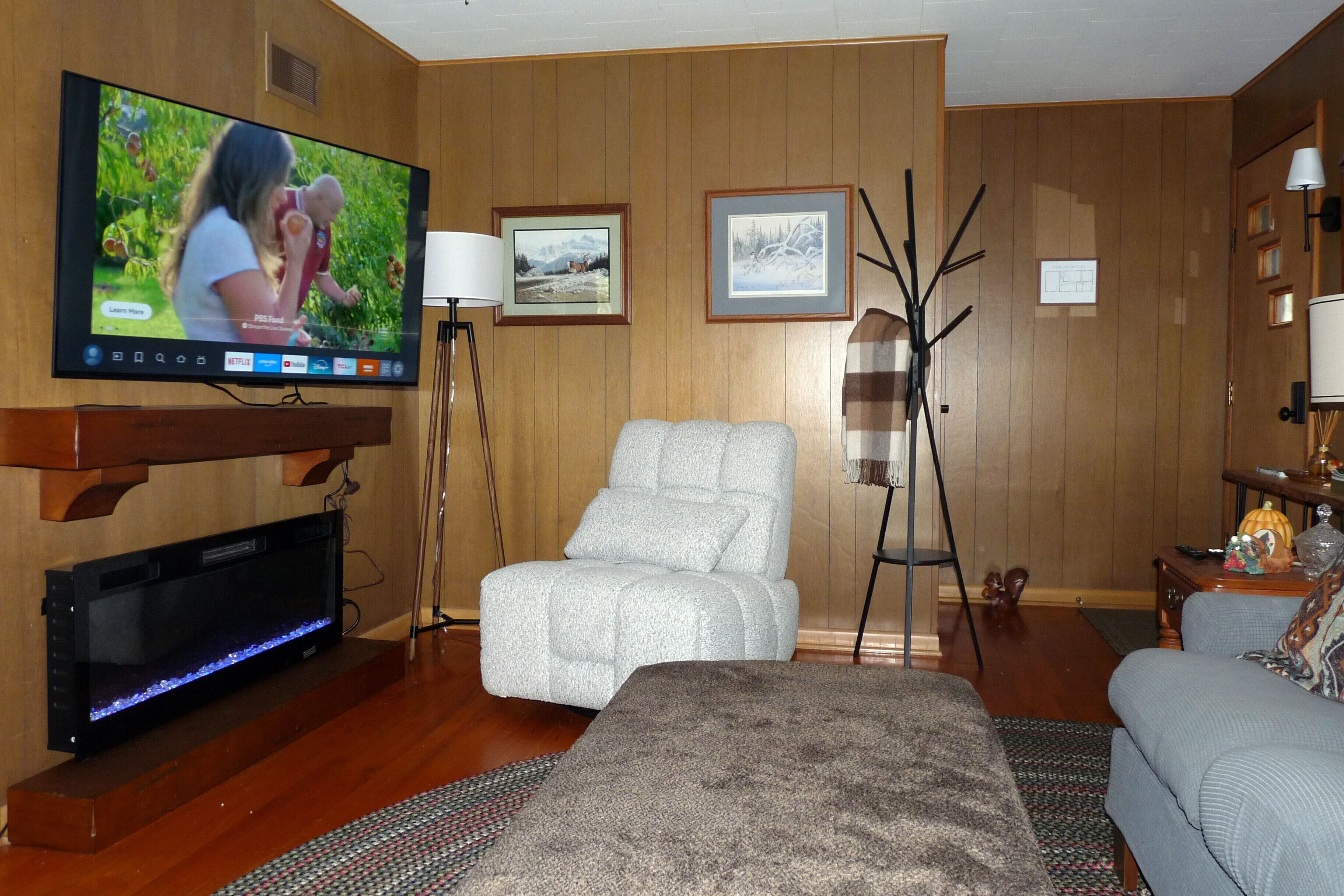 A room with a flat-screen TV mounted on the wall, a sofa, a chair, and a small table with a lamp.