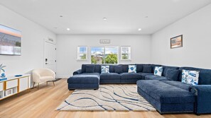 Una sala de estar moderna con un sofá modular, un sillón blanco, una mesa de centro y una alfombra. Hay ventanas con persianas, una puerta y un televisor montado en la pared.