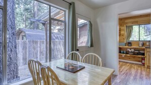 A dining area with a wooden table and chairs, a view of a wooden cabin through large windows, and a kitchen area with wooden cabinets and a coffee maker. - Cozy South Lake Tahoe Cabin | Near Beaches, Mountain Bike Trails & Heavenly (South Lake Tahoe)