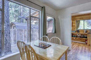 Ein Essbereich mit Holztisch und Stühlen, Blick auf eine Holzhütte durch große Fenster sowie eine Küchenzeile mit Holzschränken und einer Kaffeemaschine.