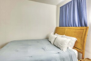 Ein Einzelbett mit Holz Kopfteil, weißen Kissen und einer hellblauen Bettdecke in einem Zimmer mit schlichten Wänden und einem Fenster mit blauen Vorhängen.