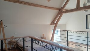 Een slaapkamer met schuine wand, houten balken, een bed met witte dekbedovertrek, een klein nachtkastje en een hanglamp.