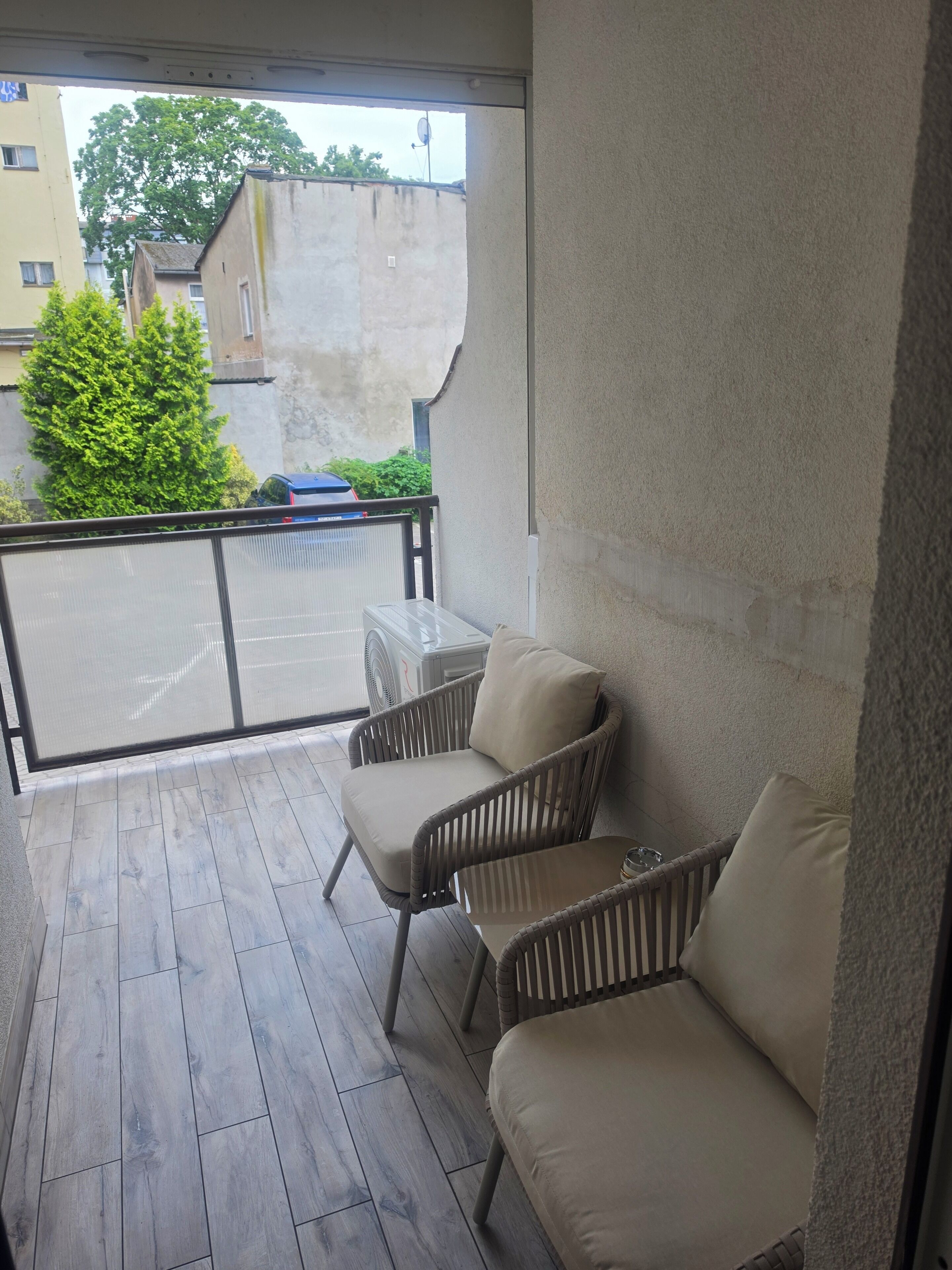 Een balkon met twee rieten stoelen en een kleine tafel, uitzicht op de straat, en een gebouw met een schotelantenne.