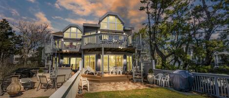 A modern two-story house with a covered patio, outdoor seating, and a large window overlooking the yard.