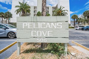 Schild für 'PELICANS COVE' mit Blick auf einen Parkplatz und Gebäude im Hintergrund.