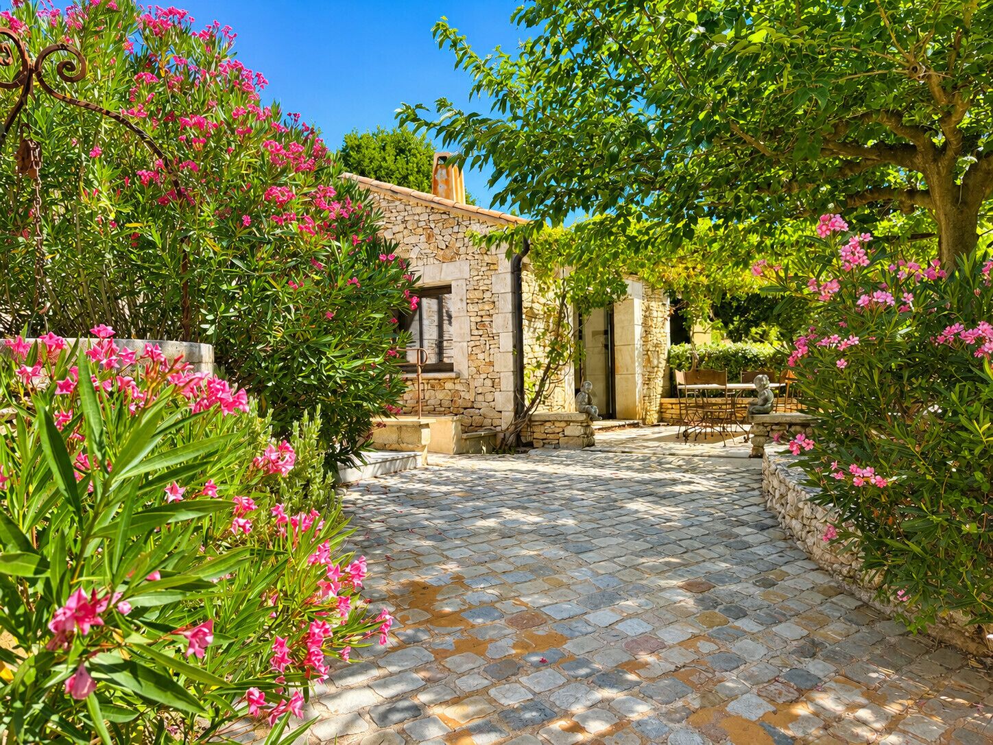 Un sentier de pierres mène à un bâtiment rustique en pierre, doté d’une fenêtre et d’une porte, et entouré d’une végétation luxuriante et de fleurs roses.