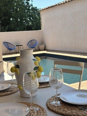 Une table à manger dressée pour deux personnes, avec un vase et des verres, une piscine en arrière-plan et des chaises bleues.