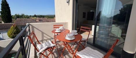 Un balcón con mesa lista para comer, sillas y un ventanal que da a un espacio interior.