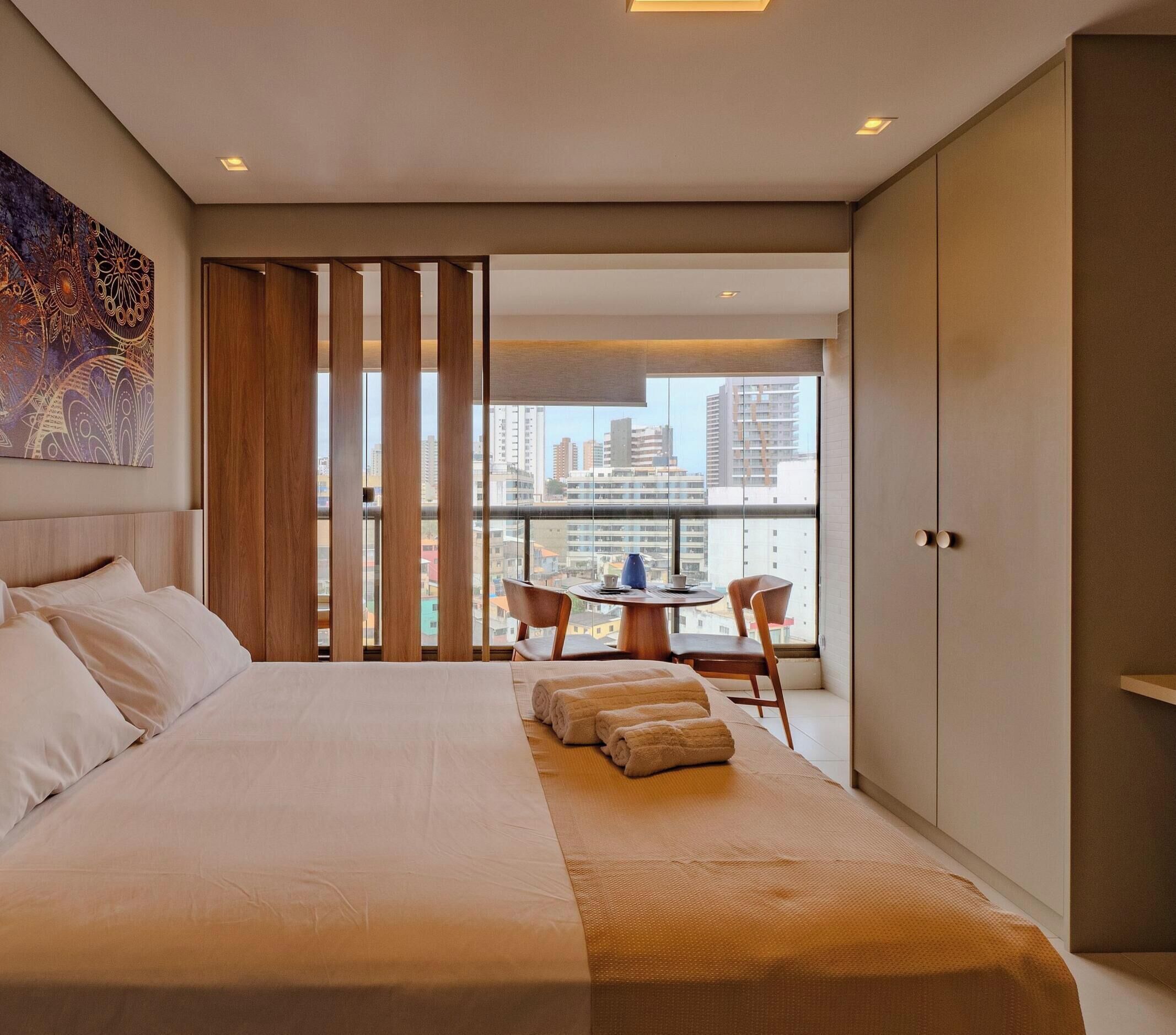 Une chambre d’hôtel moderne dotée d’un grand lit, d’un coin repas avec une table et des chaises, et offrant une vue sur le paysage urbain grâce à de grandes fenêtres.