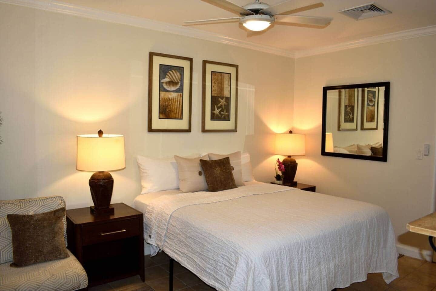 Habitación de hotel con cama, dos lámparas de noche, ventilador de techo y cuadros en las paredes.