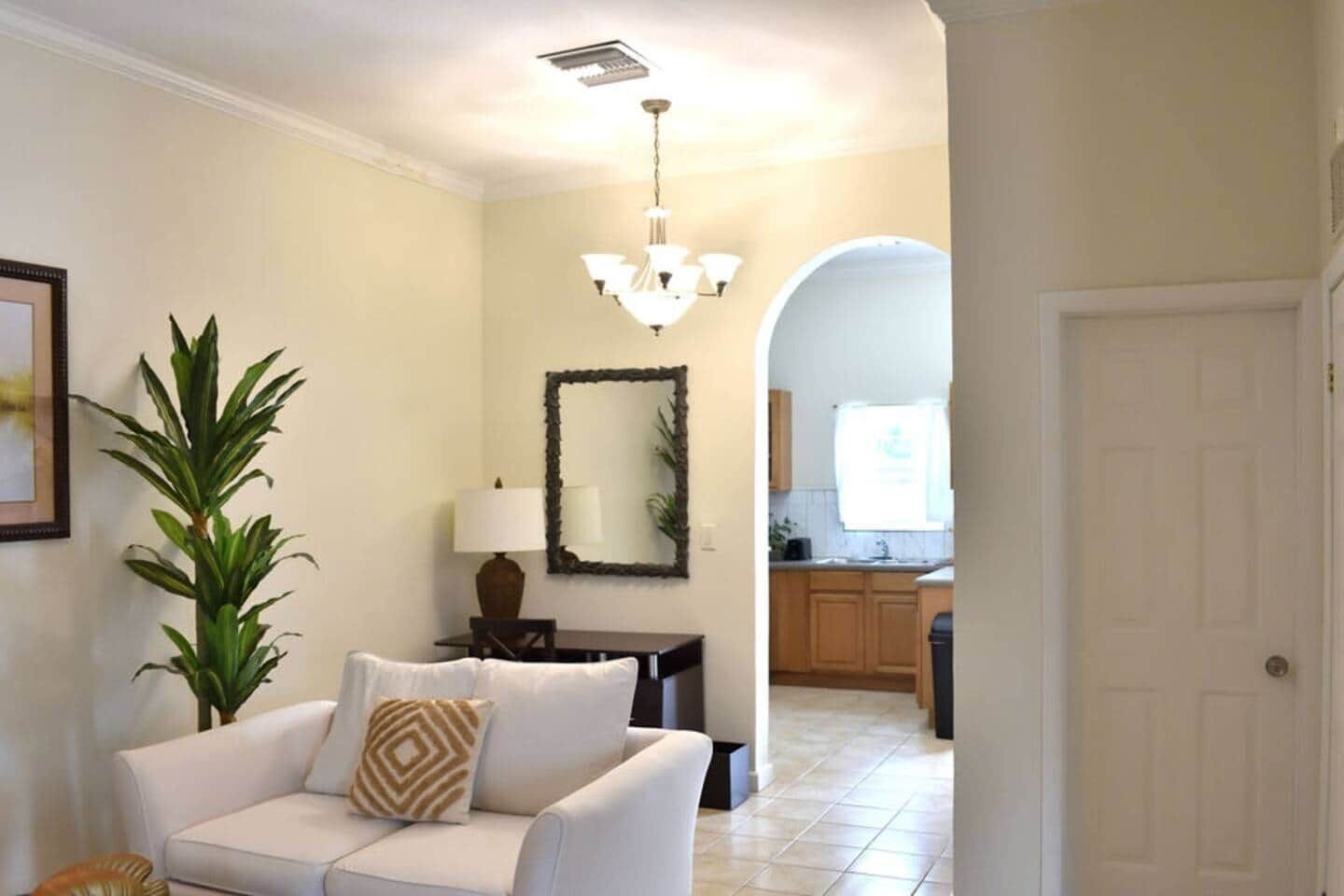A living room with a white sofa, a potted plant, a chandelier, and a kitchen visible through an archway.