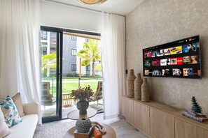 A modern living room with a large flat-screen TV mounted on the wall, a sofa, a coffee table with decorative items, and a sliding glass door leading to an outdoor area with a view of a building and palm trees. - First-floor apartment with pool view (Punta Cana)