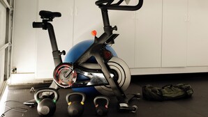 Ein stehendes Fahrrad, Kettlebells und ein blauer Trainingsball in einem Raum mit weißen Wänden und einer Tür.