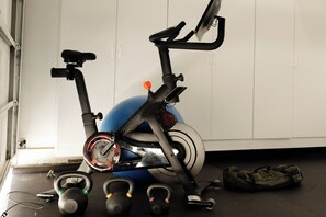 Ein stehendes Fahrrad, Kettlebells und ein blauer Trainingsball in einem Raum mit weißen Wänden und einer Tür.