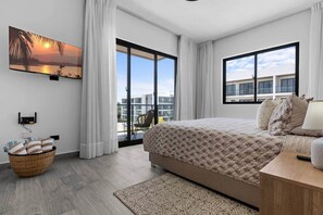 A modern bedroom with a large bed, a view of the city through the window, and a balcony with a chair. - New Luxury Condo | Pool View | Minutes to Beach (Punta Cana)